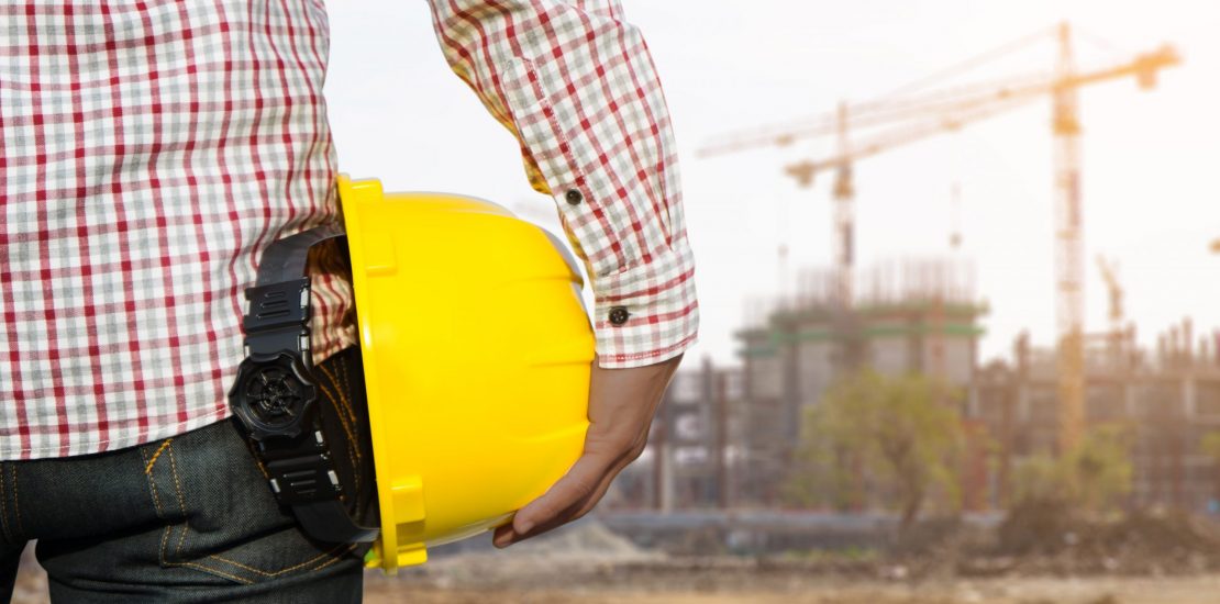 Hand's engineer worker holding yellow safety helmet with building on site background.